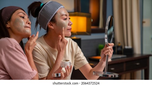 Smiling Asian Woman Friends Sitting On The Bed With Applying Skin Care Facial Mask On Their Face Together At Home. Female Gay Couple Relax And Enjoy Beauty Facial Treatment Together With Happiness