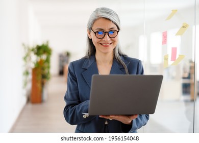 The Smiling Asian Woman In Glasses And A Suit Holding A Laptop In Her Hands While Standing In A Bright Office