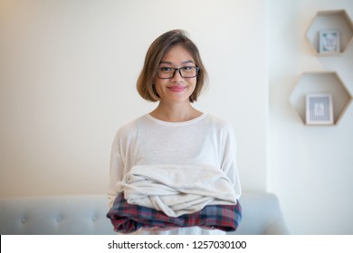 Smiling Asian Woman Holding Clean Folded Clothes At Home. Pretty Young Lady Looking At Camera And Standing With Sofa In Background. Laundry And Household Concept. Front View.