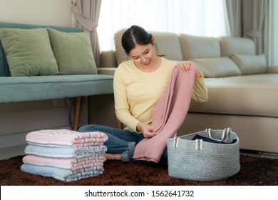 Smiling Asian Woman Holding Clean Folded Clothes At Home. Pretty Young Lady Sitting In Floor With Sofa In Background. Laundry And Household Concept. Front View.