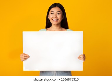 Smiling Asian Woman Holding Empty White Poster Advertising Something Standing On Yellow Studio Background. Mockup