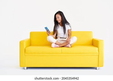 Smiling Asian Woman Holding Smartphone Browses Internet And Scrolls News Sitting On Yellow Couch On White Background. Technology, Communication Concept