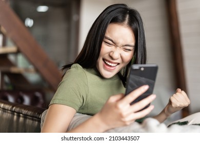 Smiling Asian Woman Looking Satisfied, Making Fist Pump And Reading Mobile Phone, Sitting At Home And Triumphing