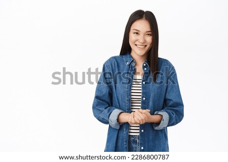 Smiling asian woman looks ready to help, friendly smile, hands clasped together, polite pose, stands over white background.