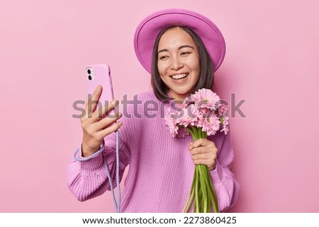 Smiling Asian woman makes selfie via smartphone wears hat and knitted sweater smiles pleasantly holds bouquet of gerbera flowers isolated over pink background. Women and spring time concept.