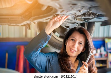 Smiling Asian Woman Mechanic Holding Wrench Standing Under A Vehicle At The Repair Garage. Female Working Underneath A Lifted Car.