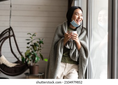 Smiling Asian Woman In Medical Face Mask, Staying At Home On Self Quarantine During Covid 19, Being Sick, Drinking Hot Tea And Looking Outside Window With Happy Laughing Face