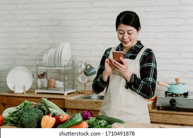 Smiling Asian Woman In Modern Kitchen Writing Text Message On Mobile Phone. Beautiful Young Lady Typing Sms Online On Cellphone During Cooking Time With Fresh Vegetables On Wooden Counter Table.