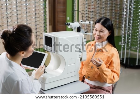 smiling asian woman pointing with hand near blurred oculist with digital tablet and vision screener in optics shop