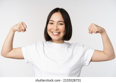 Smiling Asian Woman Showing Flexing Biceps, Muscles Strong Arms Gesture, Standing In White Tshirt Over White Background