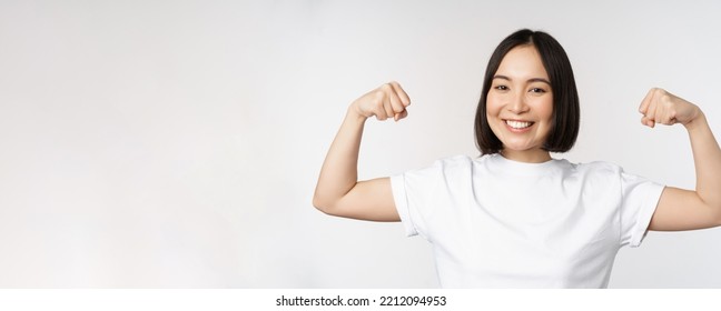 Smiling Asian Woman Showing Flexing Biceps, Muscles Strong Arms Gesture, Standing In White Tshirt Over White Background