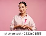 Smiling Asian woman smiling and showing pink ribbon for breast cancer awareness on pink background. Breast cancer awareness concept