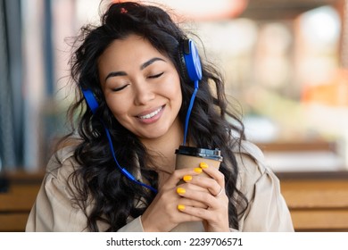 Smiling Asian woman in sidewalk cafe listening to he music via headphones while drinking coffee. Close-up portrait of a happy Asian woman with closed eyes. - Powered by Shutterstock