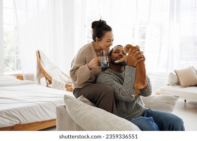 Smiling asian woman in sweater holding coffee near african american boyfriend playing with dog at home - Powered by Shutterstock