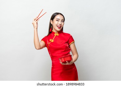 Smiling Asian woman in traditional red Chinese qipao dress holding plastic food box and chopsticks in studio isolated light gray background