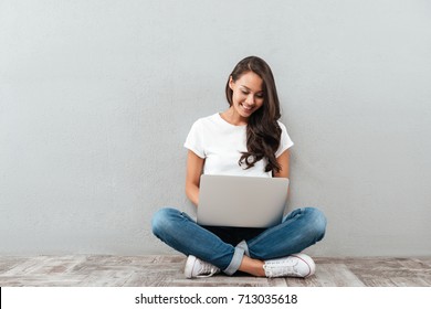 Lächelnde asiatische Frau, die während ihres Sitzens auf dem Boden auf dem Laptop einen Computer tippt und die Beine einzeln auf grauem Hintergrund gekreuzt