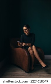 Smiling asian woman using a mobile phone while sitting on a couch in her office. Happy young woman reading a text message on her smartphone. - Shutterstock ID 2224618549