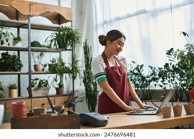 A smiling Asian woman wearing an apron works on laptop in her plant store, surrounded by greenery. - Powered by Shutterstock