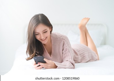 Smiling Asian woman wearing knitted sweater pink cold looking at mobile phone lying down on white bed. Beautiful girl typing on smartphone and surfing the net.Concept woman lifestyle and winter. 