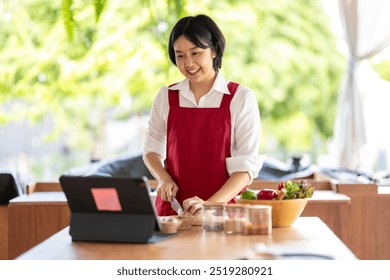 Smiling asian woman wearing a red apron is slicing vegetables while looking at a recipe on a tablet computer - Powered by Shutterstock