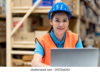 Smiling Asian Woman Worker In Safety Vest And Helmet Sitting And Working With Computer Laptop In Factory Warehouse. People, Warehouse And Industry Concept