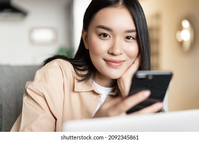 Smiling Asian Woman Working From Home On Laptop, Looking At Smartphone