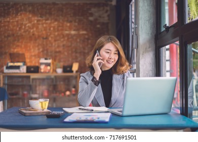 Smiling Asian Woman Working At Home Office ,Thai Young Girl Calculating Bills By Laptop Computer Sitting On Wooden Desk And Calling On Phone Land Line And Looking At Laptop Screen In The Living Room 