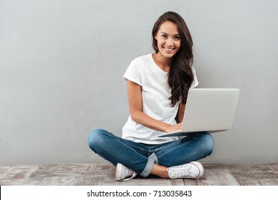 Smiling asian woman working on laptop computer while sitting on the floor with legs crossed isolated over gray background