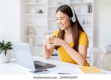 Smiling asian woman in a yellow top enjoying coffee while on a video call at her workspace, using laptop and headphones - Powered by Shutterstock