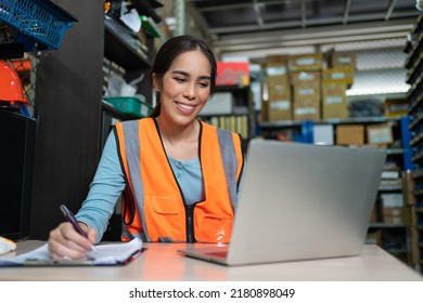 Smiling Asian young female sitting and using laptop at warehouse factory.Woman worker in vest safety writing order from customer into paper clipboard in store office. - Powered by Shutterstock