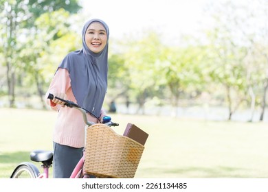 Smiling Asian young muslim woman wear hijab headscarf riding bicycle with book on basket in the park. Education and field trip outdoor concept - Powered by Shutterstock