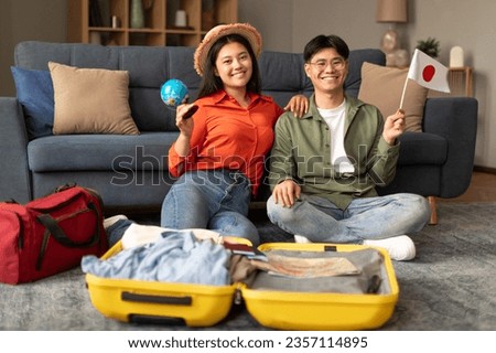 Smiling Asian Young Spouses Posing With Flag Of Japan And World Globe, Sitting Near Opened Suitcase, Planning Their Vacation Travel Or Relocation To New Country At Home Interior, Selective Focus