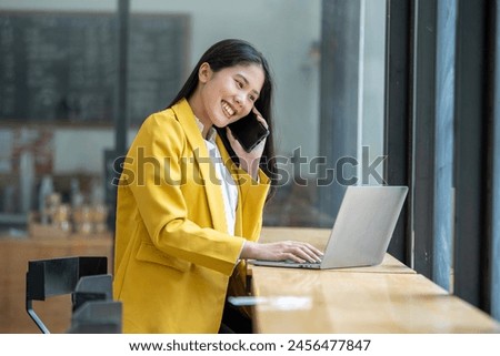 Smiling Asian young woman enjoys using her smartphone while sitting at a modern work desk with a laptop.

