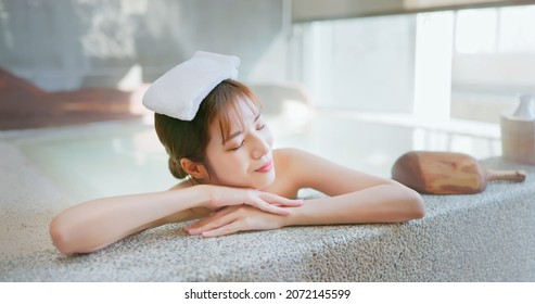 smiling asian young woman lying on the bathtub is relaxing in hot spring with towel on head