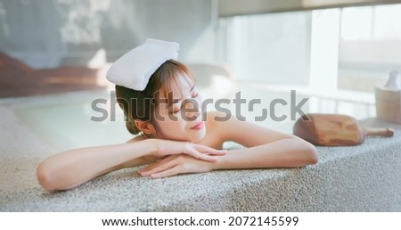 smiling asian young woman lying on the bathtub is relaxing in hot spring with towel on head