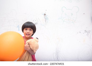 A Smiling Asian Youngster Carrying An Orange Balloon While Donning A Pink Outfit. On The House's White Wall, He Painted With Several Crayons. Copy Space.