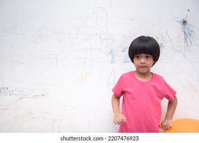 A Smiling Asian Youngster Carrying An Orange Balloon While Donning A Pink Outfit. On The House's White Wall, He Painted With Several Crayons. Copy Space.