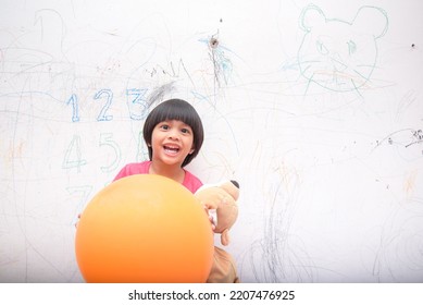 A Smiling Asian Youngster Carrying An Orange Balloon While Donning A Pink Outfit. On The House's White Wall, He Painted With Several Crayons. Copy Space.