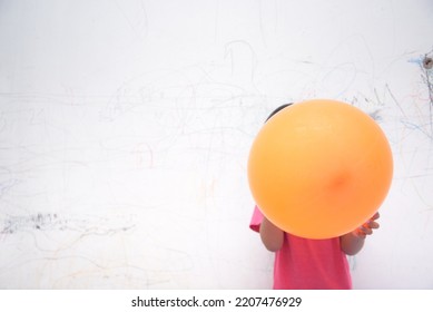 A Smiling Asian Youngster Carrying An Orange Balloon While Donning A Pink Outfit. On The House's White Wall, He Painted With Several Crayons. Copy Space.