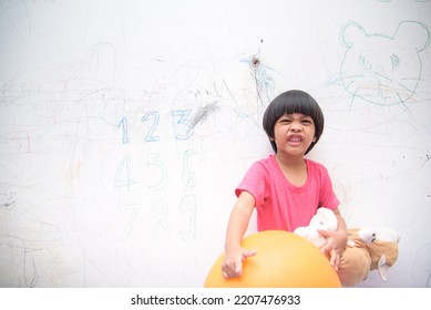 A Smiling Asian Youngster Carrying An Orange Balloon While Donning A Pink Outfit. On The House's White Wall, He Painted With Several Crayons. Copy Space.