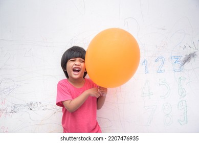 A Smiling Asian Youngster Carrying An Orange Balloon While Donning A Pink Outfit. On The House's White Wall, He Painted With Several Crayons. Copy Space.