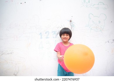 A Smiling Asian Youngster Carrying An Orange Balloon While Donning A Pink Outfit. On The House's White Wall, He Painted With Several Crayons. Copy Space.