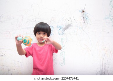 A Smiling Asian Youngster Carrying An Orange Balloon While Donning A Pink Outfit. On The House's White Wall, He Painted With Several Crayons. Copy Space.