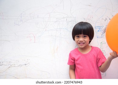 A Smiling Asian Youngster Carrying An Orange Balloon While Donning A Pink Outfit. On The House's White Wall, He Painted With Several Crayons. Copy Space.