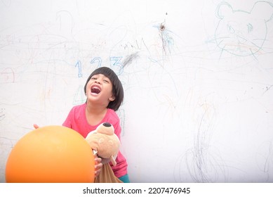 A Smiling Asian Youngster Carrying An Orange Balloon While Donning A Pink Outfit. On The House's White Wall, He Painted With Several Crayons. Copy Space.