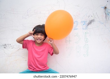 A Smiling Asian Youngster Carrying An Orange Balloon While Donning A Pink Outfit. On The House's White Wall, He Painted With Several Crayons. Copy Space.