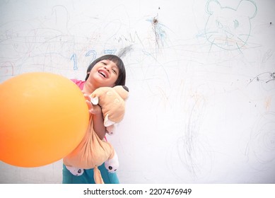 A Smiling Asian Youngster Carrying An Orange Balloon While Donning A Pink Outfit. On The House's White Wall, He Painted With Several Crayons. Copy Space.