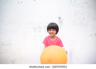 A Smiling Asian Youngster Carrying An Orange Balloon While Donning A Pink Outfit. On The House's White Wall, He Painted With Several Crayons. Copy Space.