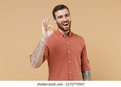 Smiling Assured Happy Young Brunet Man 20s Short Haircut With Earrings Wears Orange Shirt Showing Okay Ok Gesture Isolated On Plain Pastel Light Beige Background Studio Portrait. Tattoo Translate Fun