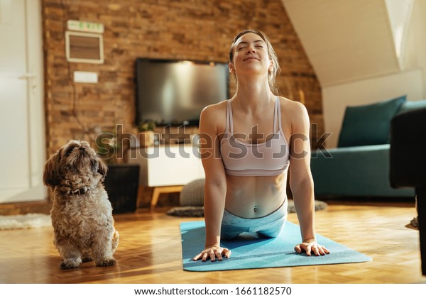 Smiling athletic woman in cobra pose practicing Yoga\
with her dog at home. 